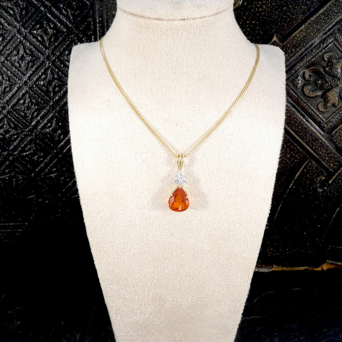 Gold necklace with a teardrop-shaped orange pendant on a mannequin bust against a textured black background.