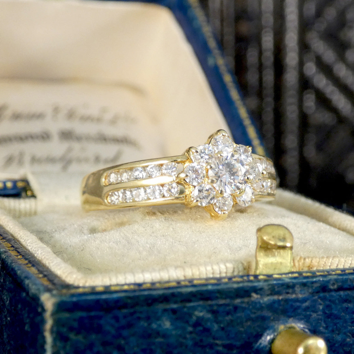 Vintage-style diamond cluster ring in 18ct yellow gold, set with a central round diamond surrounded by a floral halo and diamond-accented split shoulders, presented in a classic blue velvet ring box.