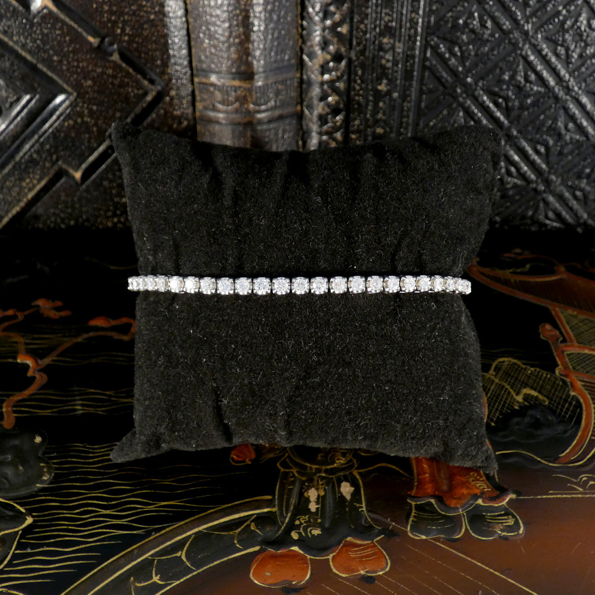 Elegant diamond tennis bracelet displayed on a black velvet cushion, featuring a continuous row of sparkling round diamonds in white gold, set against a decorative background.