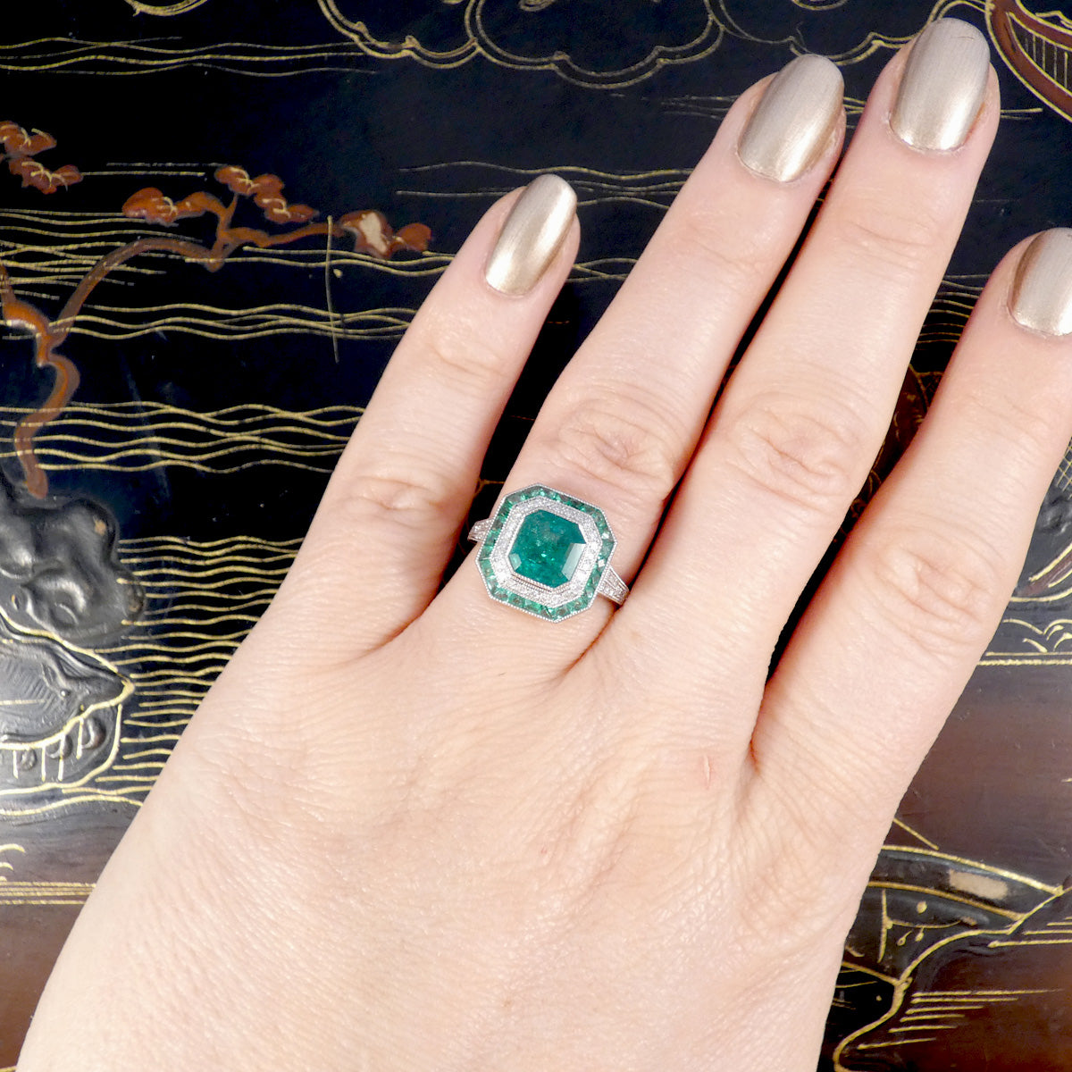 Striking Art Deco-style ring featuring a central emerald-cut emerald, surrounded by a double halo of calibre-cut emeralds and pavé-set diamonds, all set in platinum. Shown on the hand to see size and colour ratio.