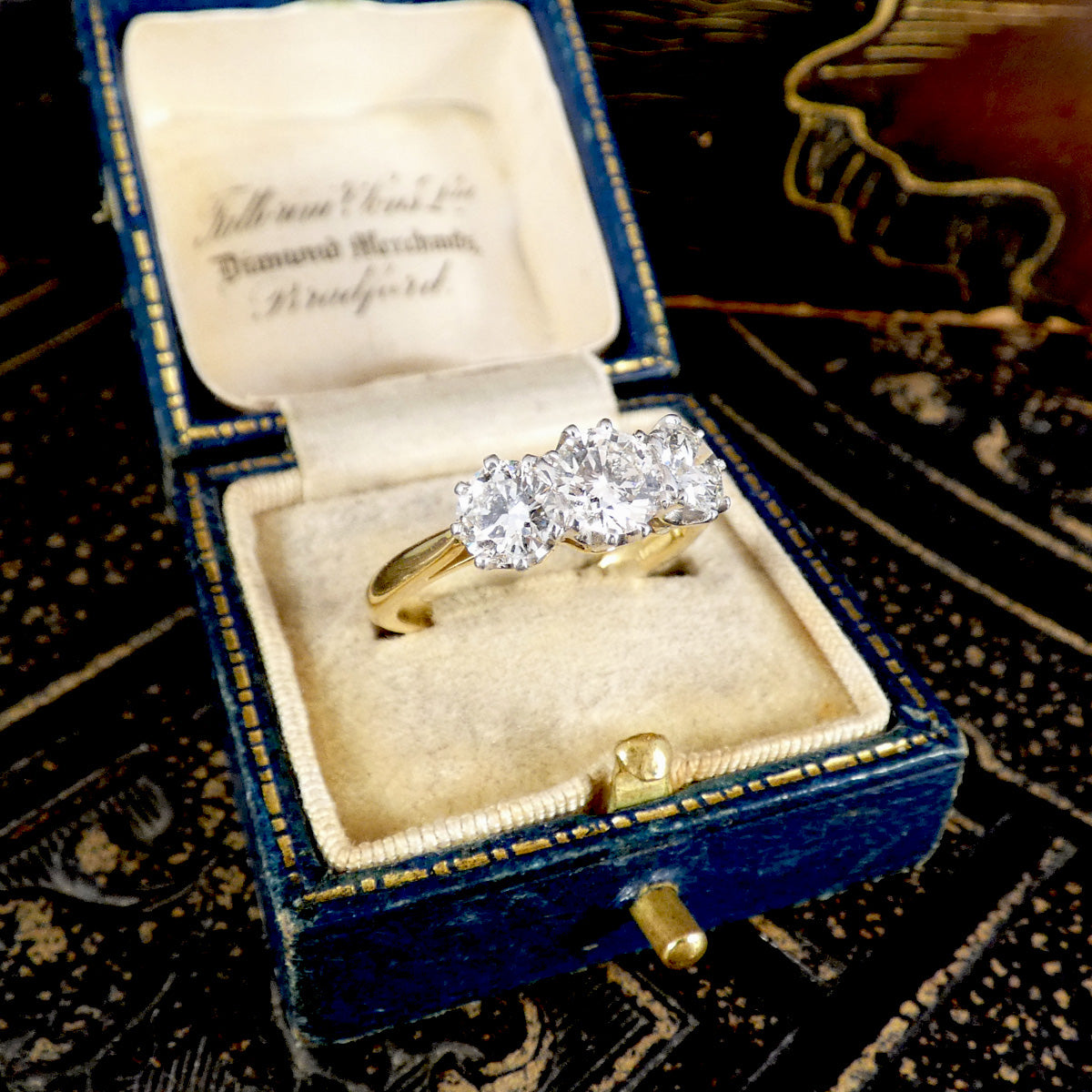 A yellow gold three-stone diamond ring displayed inside an open vintage blue velvet ring box with a cream velvet interior. The ring box lid is open, showing the brand name 'J.