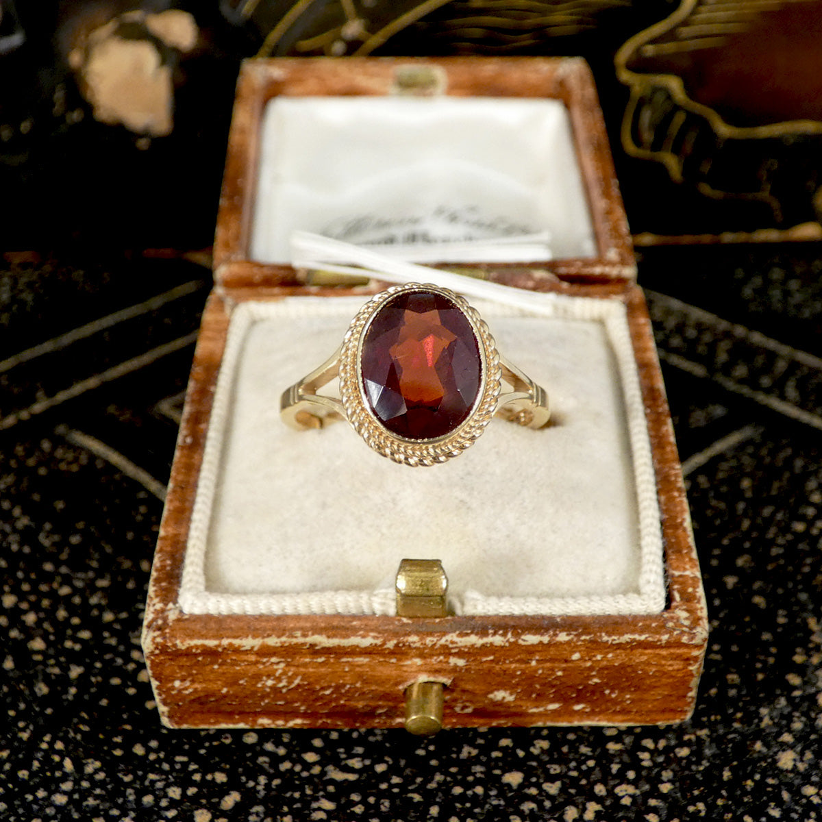 Gold ring with a red garnet gemstone in a wooden box on a dark textured surface