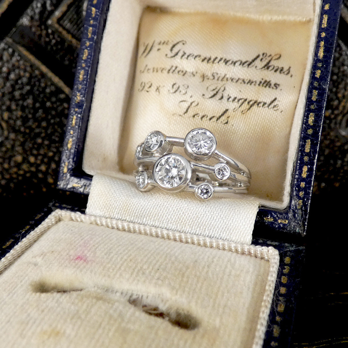A silver-toned multi-stone ring with round-cut diamonds displayed inside an open vintage jewelry box. The cream satin lining shows the brand name 'W.