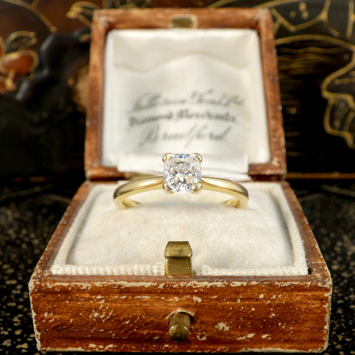 18-carat yellow gold solitaire engagement ring featuring a GIA-certified modified cushion-cut diamond of E colour, elegantly presented in a vintage-style jewellery box. The sparkling diamond and classic design make it a perfect choice for a romantic proposal.