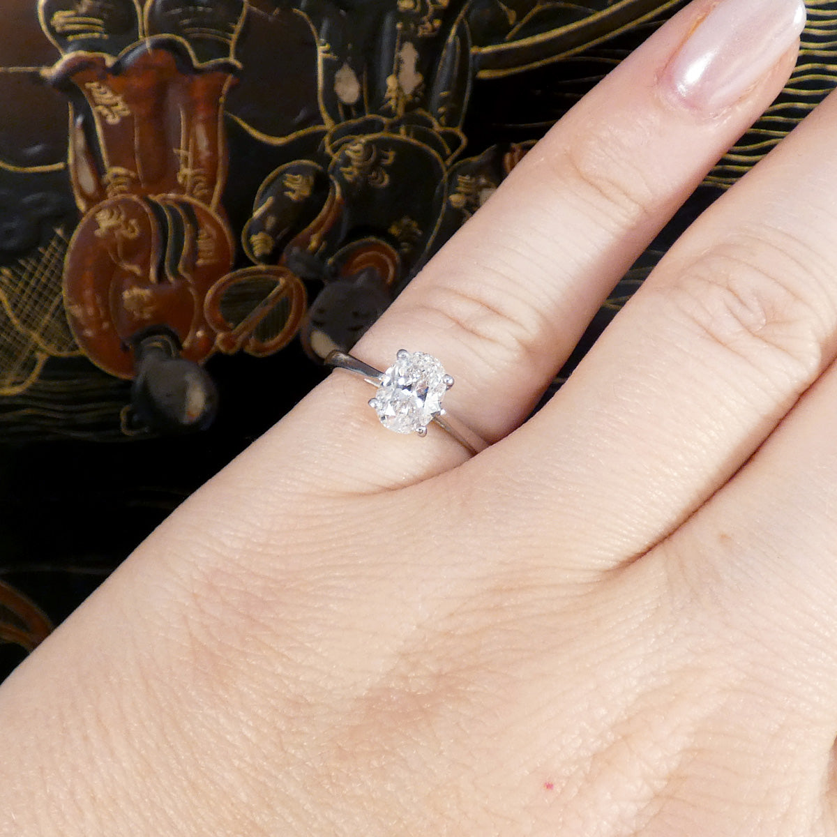 A close-up of a hand wearing a classic solitaire diamond engagement ring on a silver band, set against a dark patterned background.
