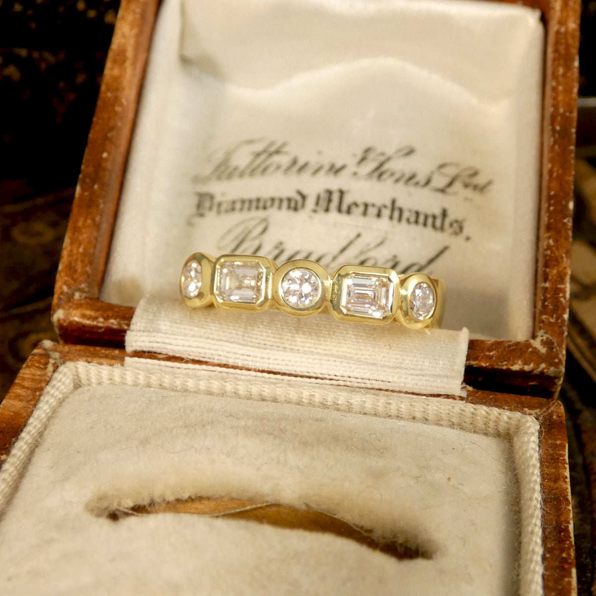 Half eternity ring with bezel-set emerald-cut and brilliant-cut diamonds in 18ct yellow gold, displayed in a vintage wooden ring box with a cream fabric interior.