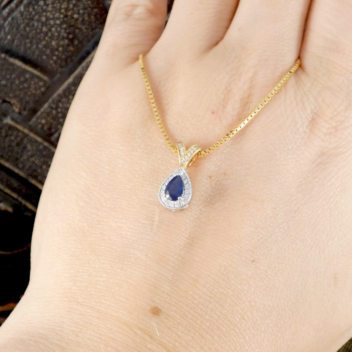 A gold chain necklace with a teardrop-shaped blue gemstone pendant surrounded by small clear stones, shown resting on a person's hand.