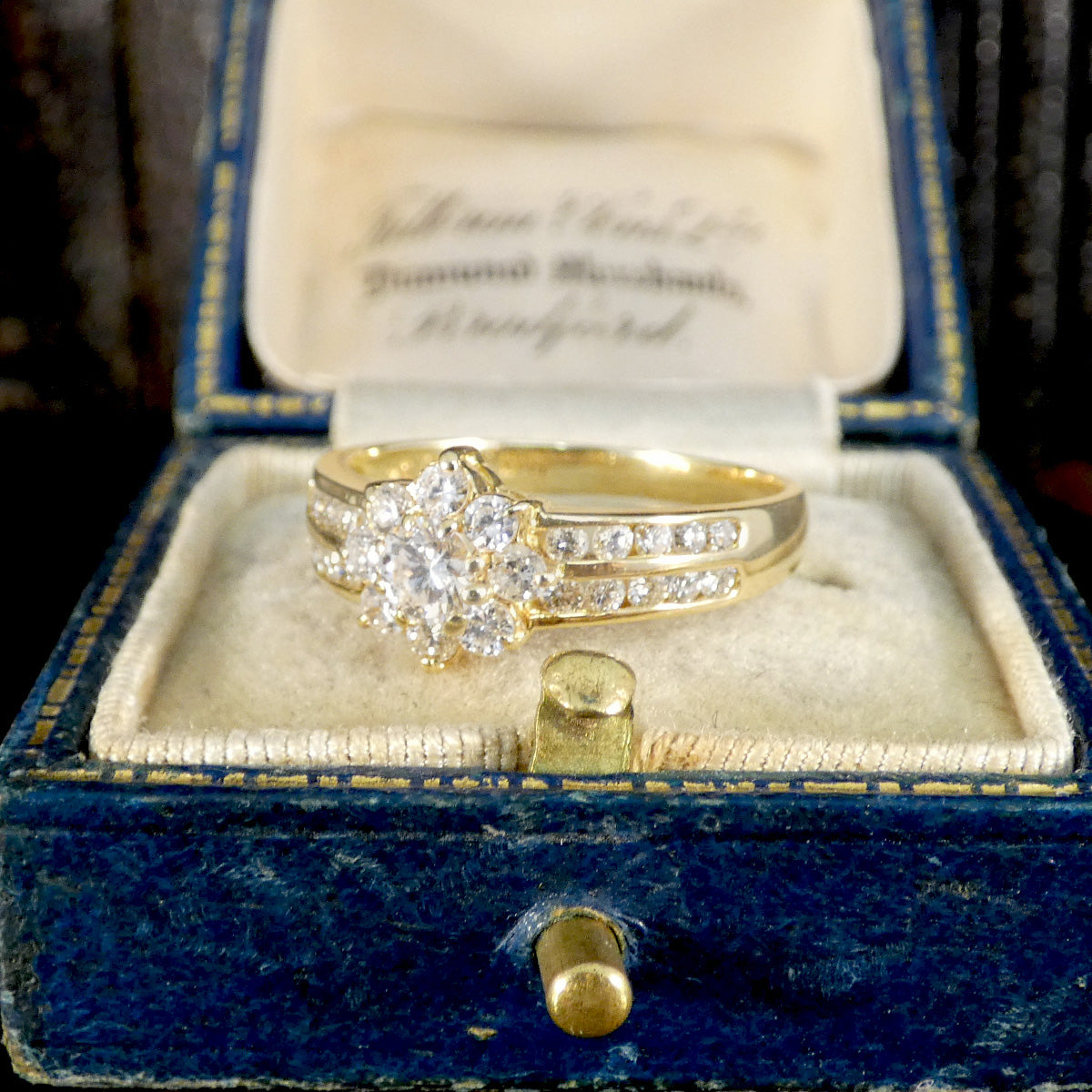 Vintage-style diamond cluster ring in 18ct yellow gold, set with a central round diamond surrounded by a floral halo and diamond-accented split shoulders, presented in a classic blue velvet ring box.