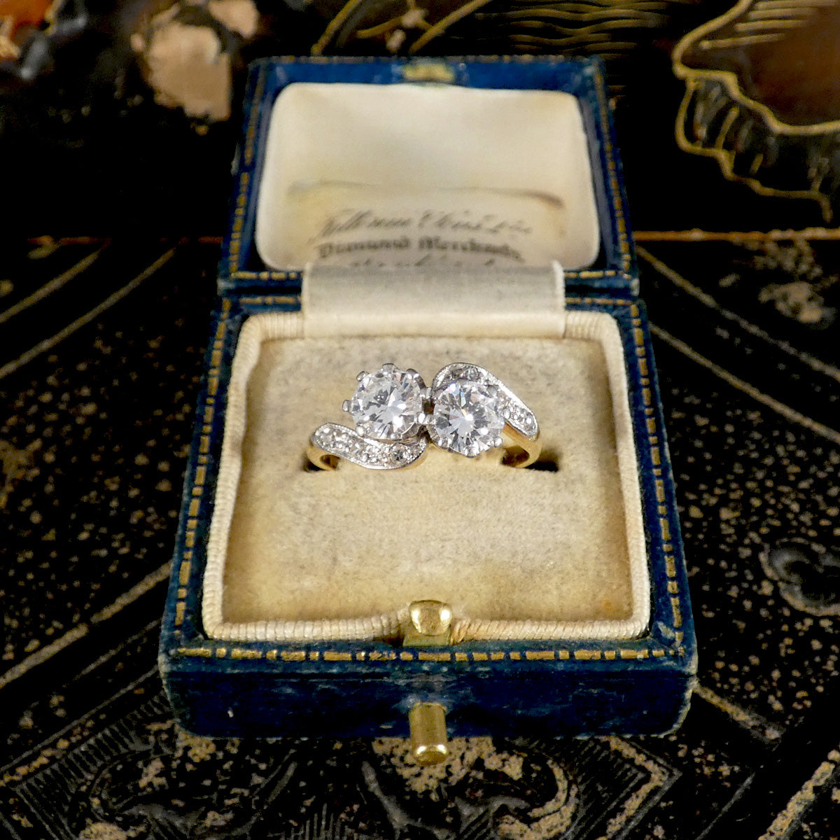 A vintage double diamond ring in a silver-toned setting, presented inside an open antique blue velvet ring box with a cream lining, set against a dark patterned background.