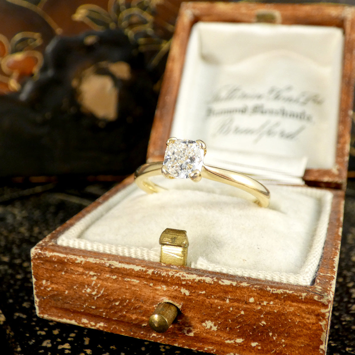 18-carat yellow gold solitaire ring featuring a GIA-certified modified cushion-cut diamond of E colour, displayed in a vintage-style wooden jewellery box with a velvet lining. The sparkling diamond and elegant design make it a standout choice for engagement or special occasions.