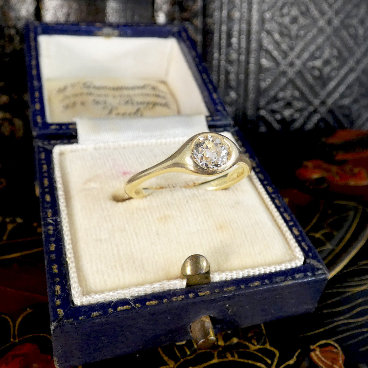A yellow gold ring with a round-cut clear gemstone displayed inside an open vintage blue velvet ring box with a cream insert.