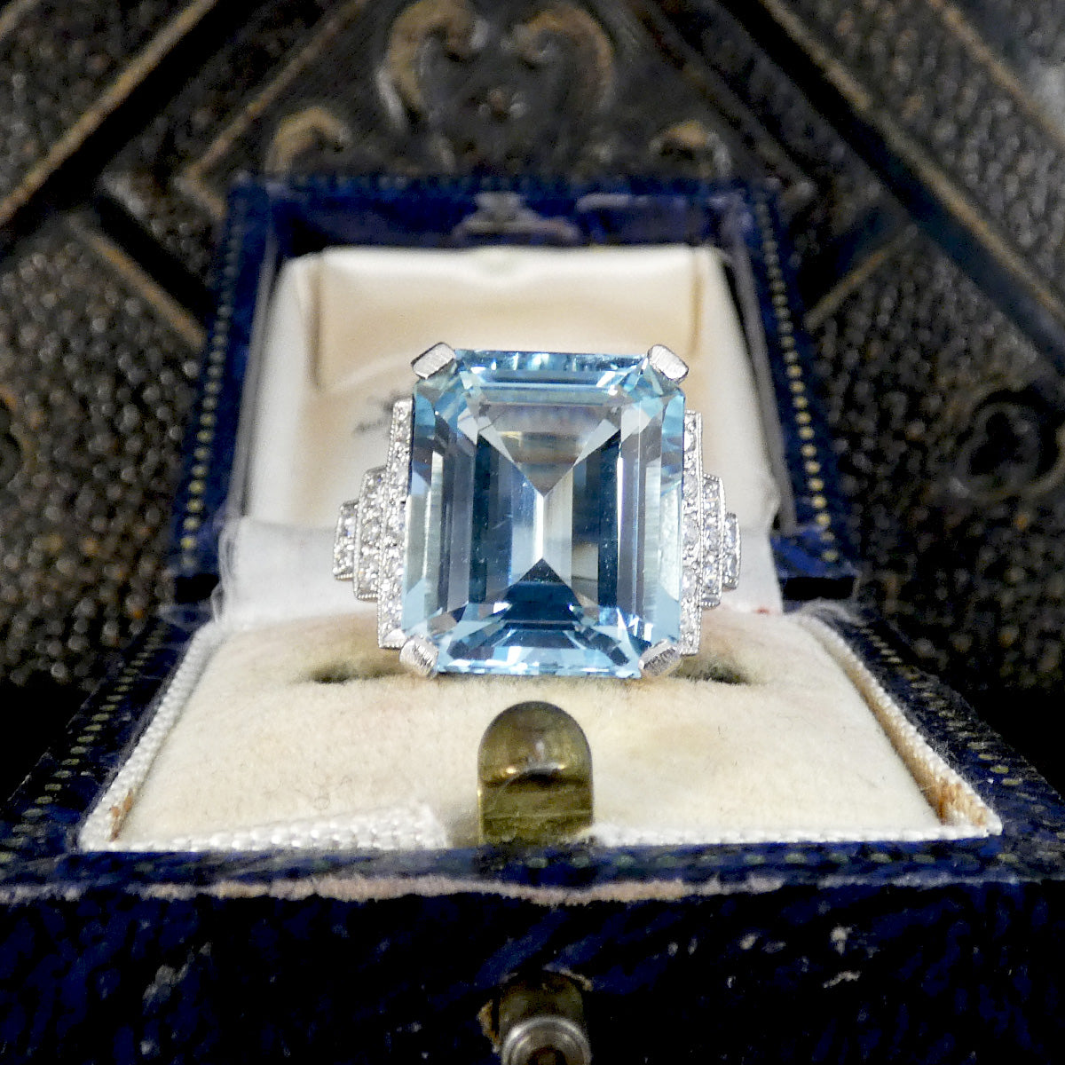 An emerald-cut light blue gemstone ring with diamond accents on a silver-toned setting, displayed in an open dark blue jewelry box with a cream cushion.
