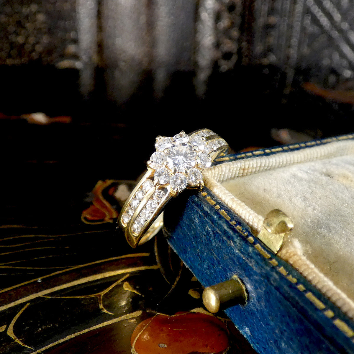 Elegant diamond cluster ring in 18ct yellow gold, featuring a floral design with a round brilliant-cut center stone and diamond-set split shoulders, displayed at an angle on a vintage blue velvet ring box.
