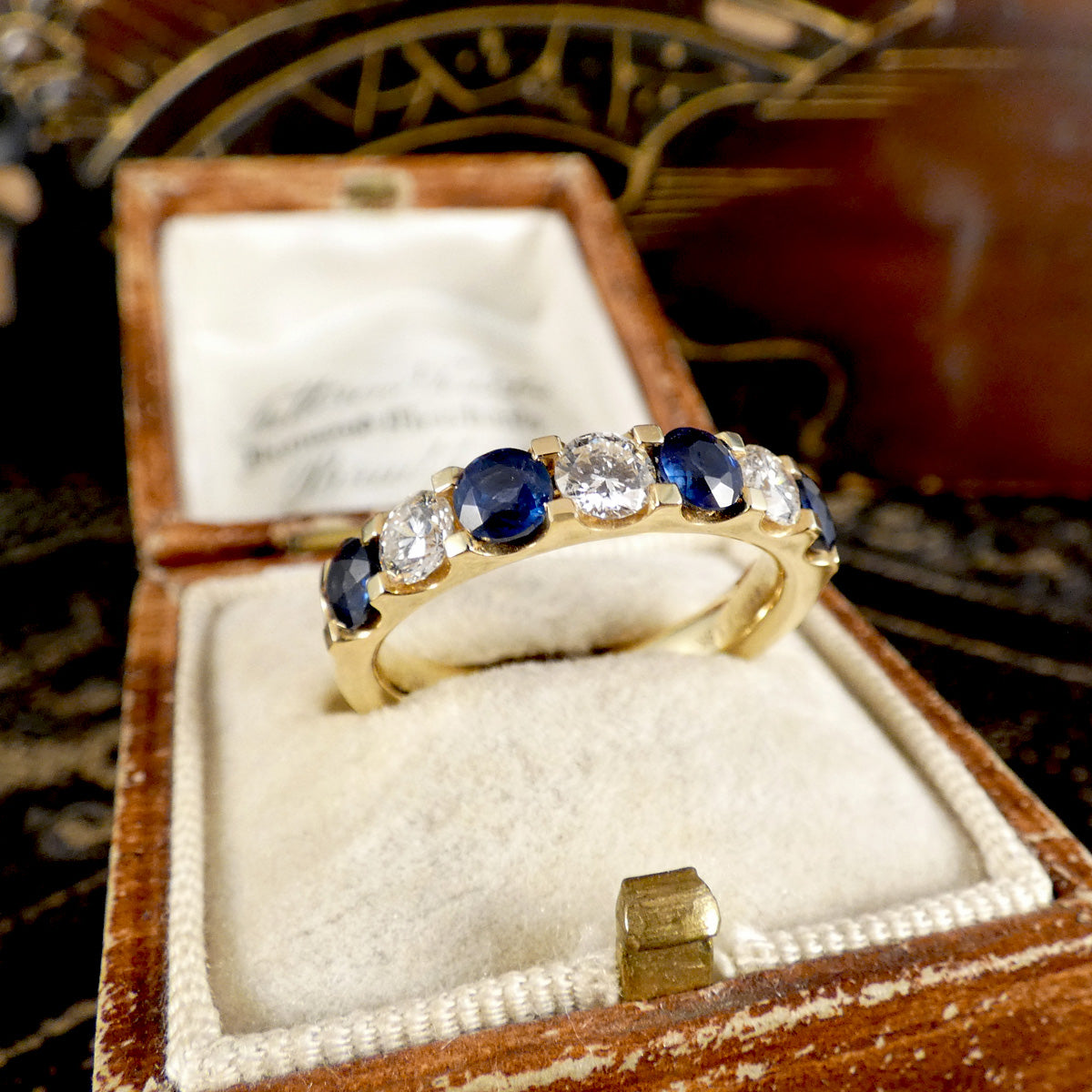 A yellow gold ring with alternating deep blue sapphires and clear round diamonds, displayed in an open vintage wooden jewelry box with a cream velvet lining.