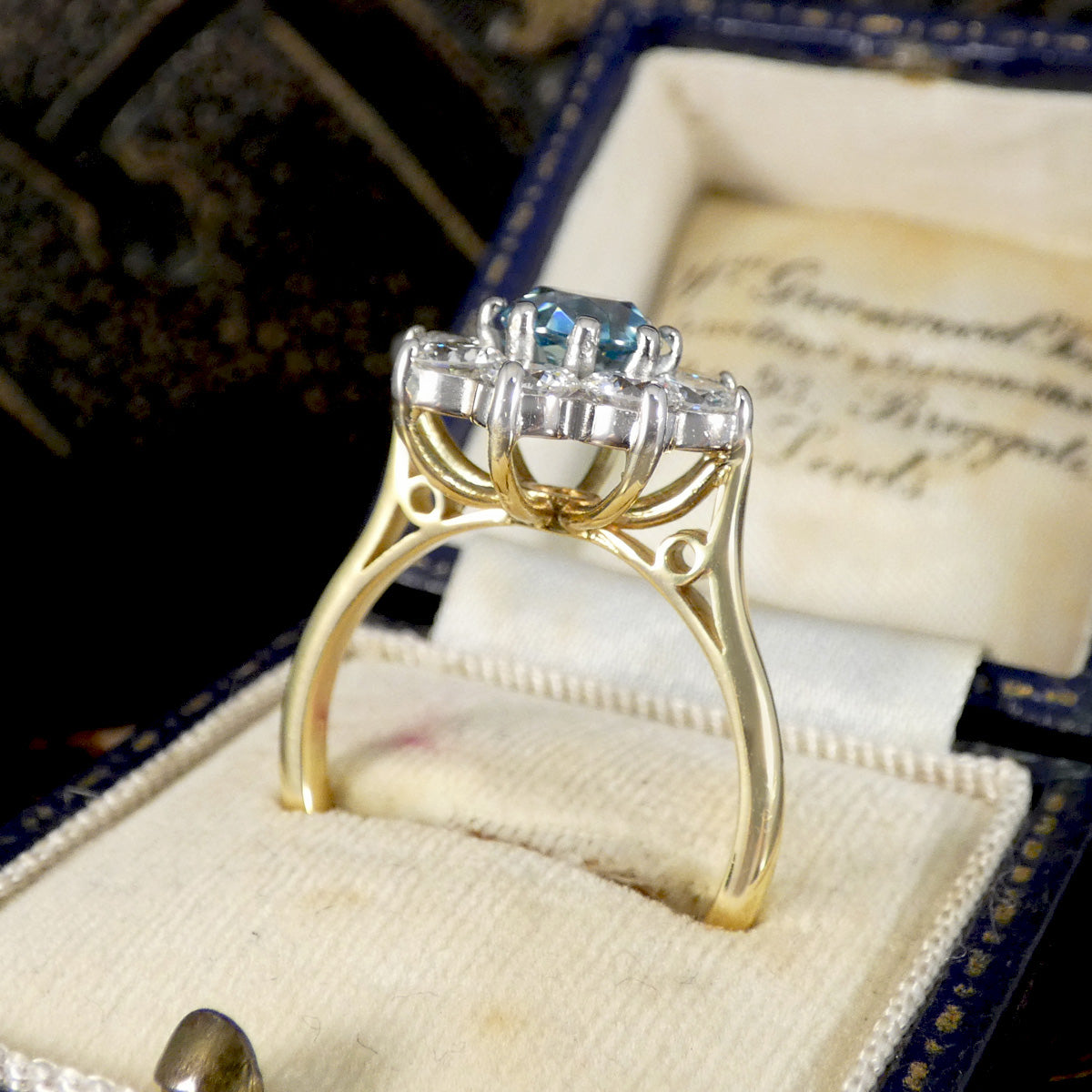 A yellow gold ring with a central round blue gemstone and surrounding clear stones, displayed inside an open vintage-style ring box with a cream cushion.