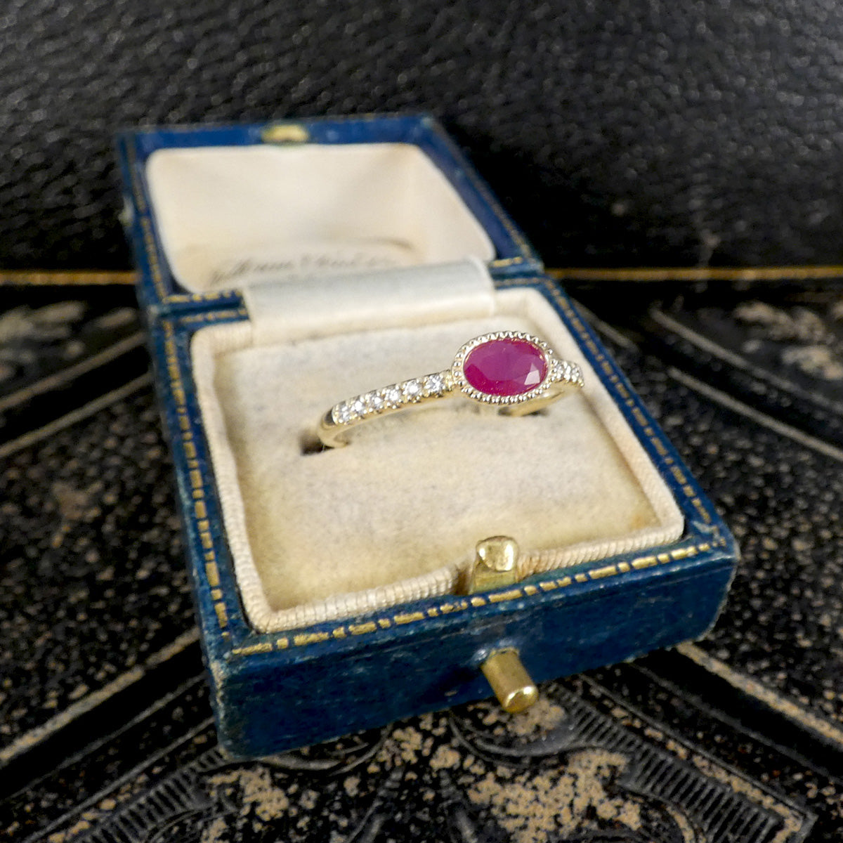 Gold ring with a red gemstone in an open blue jewelry box on a textured surface.