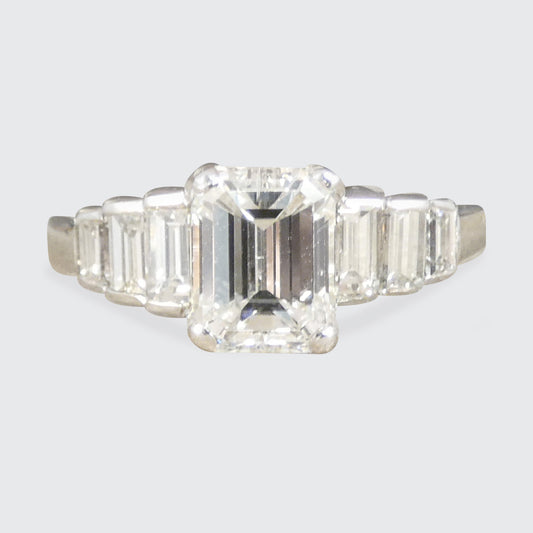 An emerald-cut diamond ring with baguette side stones in a silver-toned setting, shown against a light gray background.