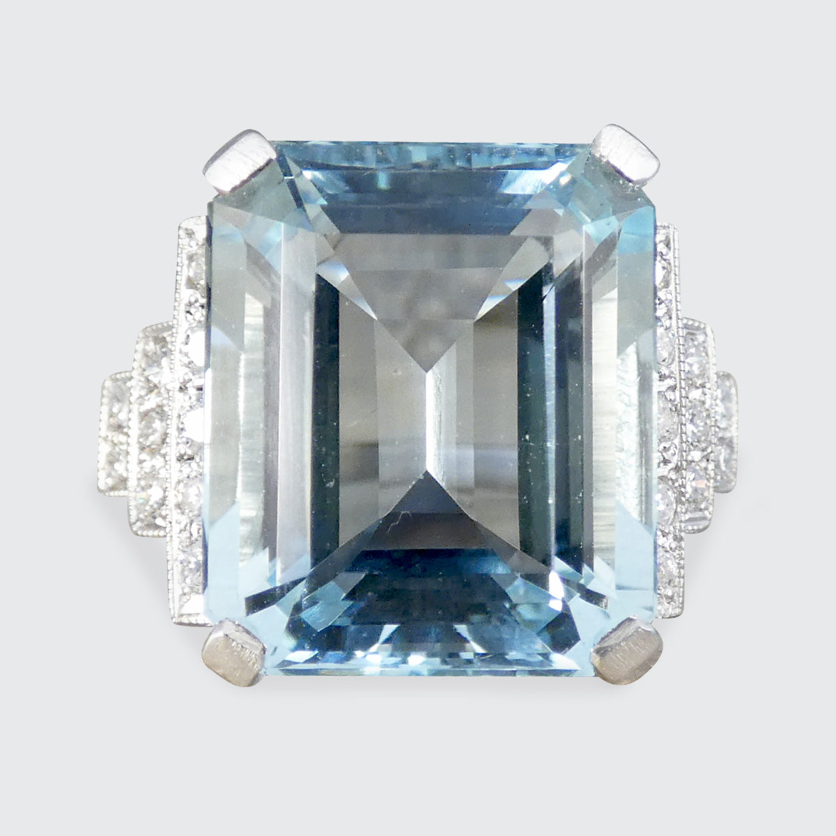 An emerald-cut light blue aquamarine ring with a silver-toned setting and small clear accent stones on the shoulders, shown against a light gray background.