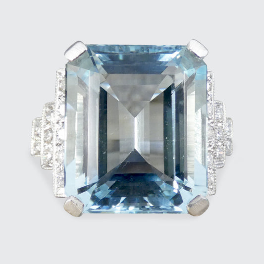 An emerald-cut light blue aquamarine ring with a silver-toned setting and small clear accent stones on the shoulders, shown against a light gray background.