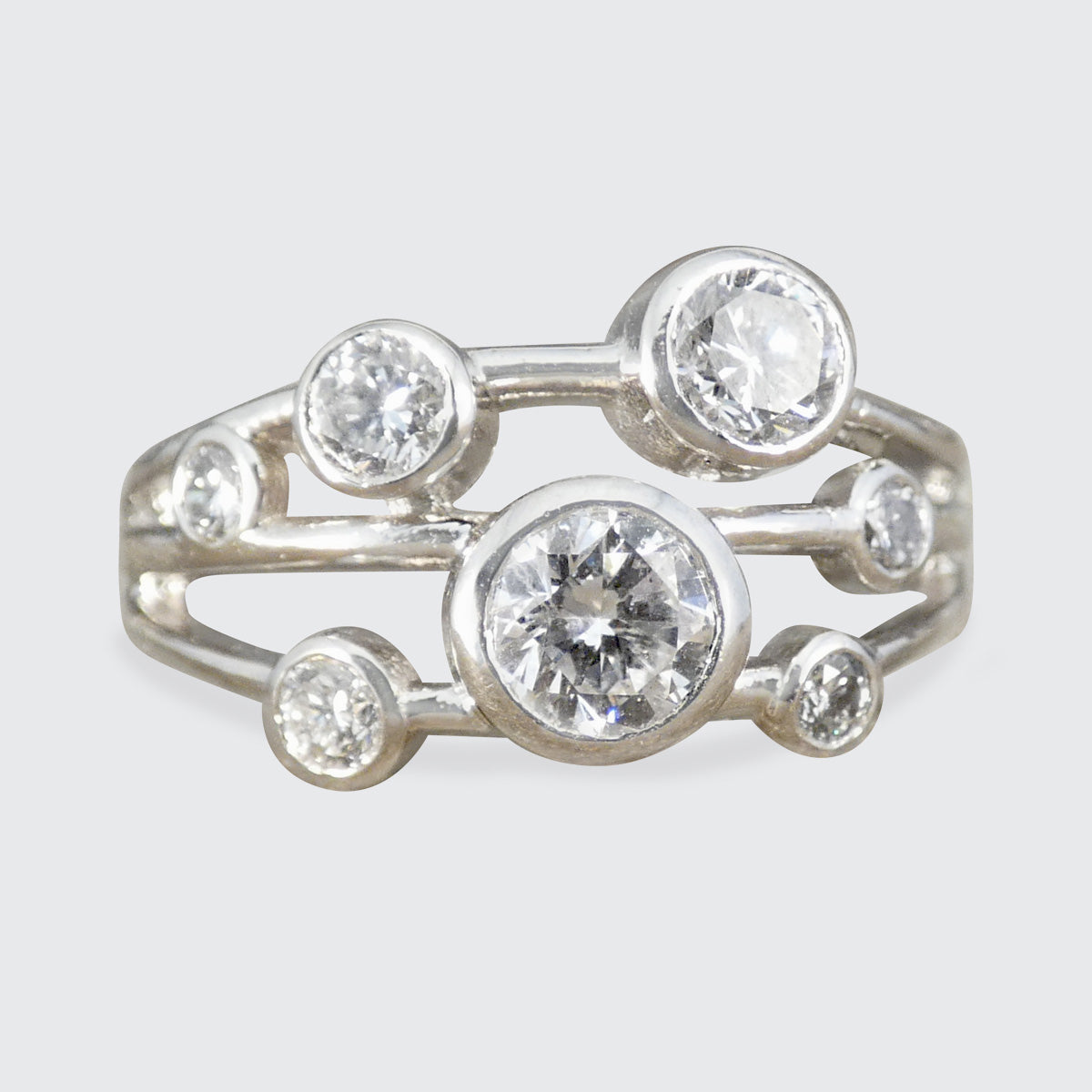 A platinum ring featuring a cluster of round clear gemstones in bezel settings, photographed against a light gray background.