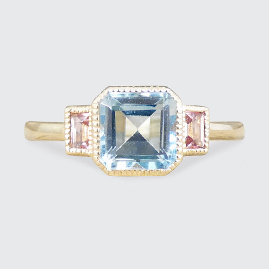 A gold ring featuring a large square-cut light blue topaz centre stone and two small rectangular pink tourmalines set on either side, shown against a light gray background.
