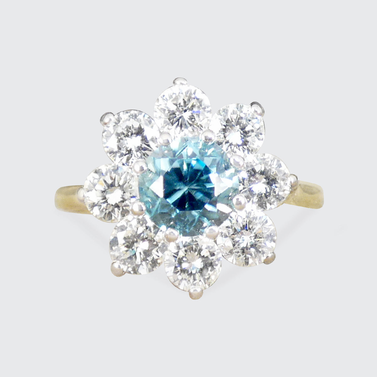 A yellow gold cluster ring featuring a central round-cut light blue gemstone surrounded by a halo of smaller clear stones, set against a plain light gray background.