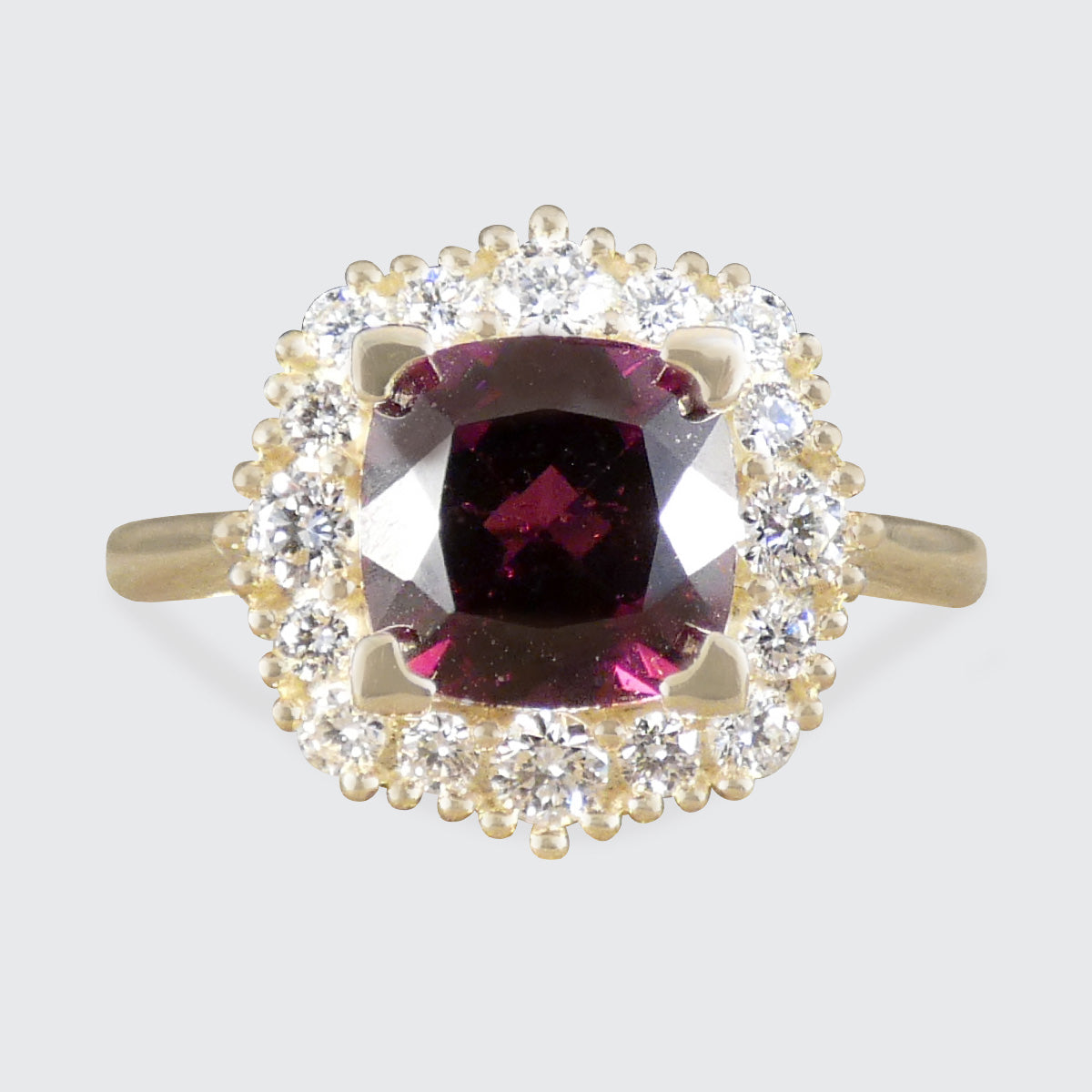 A yellow gold ring featuring a cushion-cut dark red garnet center stone surrounded by a halo of small clear round-cut stones, set against a light gray background.