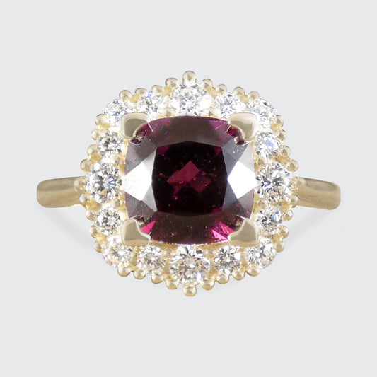 A yellow gold ring featuring a cushion-cut dark red garnet center stone surrounded by a halo of small clear round-cut stones, set against a light gray background.