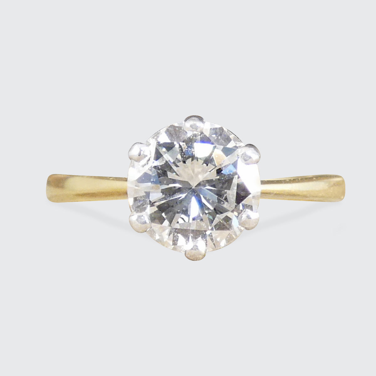 A classic solitaire engagement ring featuring a round brilliant-cut clear gemstone in a prong setting on a slender yellow gold band, shown against a neutral light gray background.