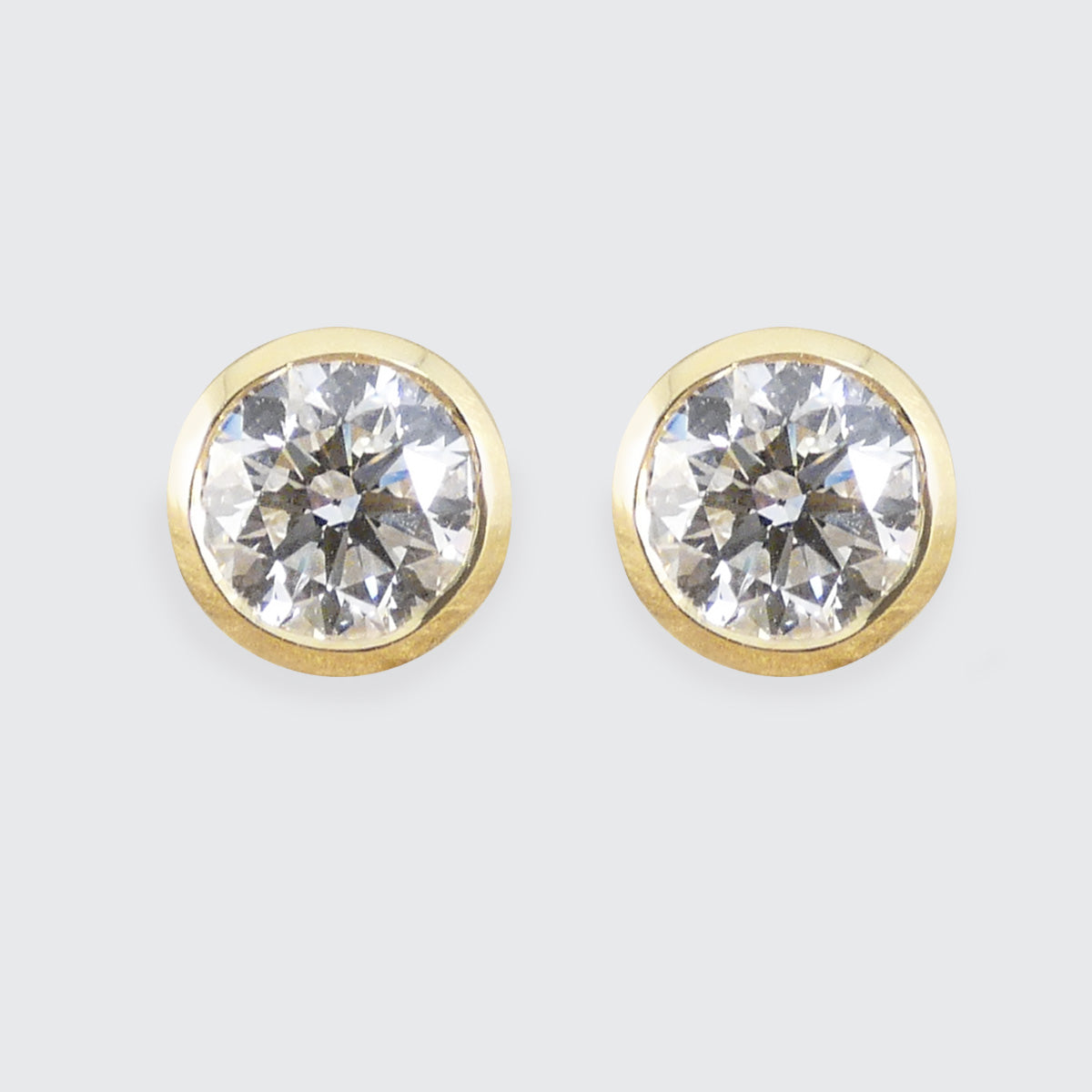 A pair of round stud earrings with clear faceted diamond gemstones in gold bezel settings, shown against a light gray background.