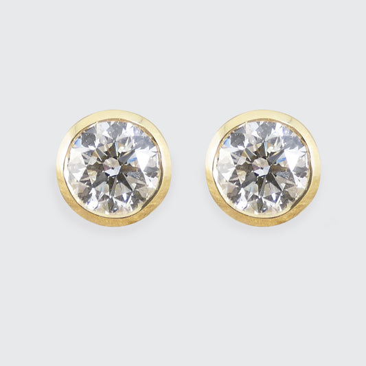 A pair of round stud earrings with clear faceted diamond gemstones in gold bezel settings, shown against a light gray background.