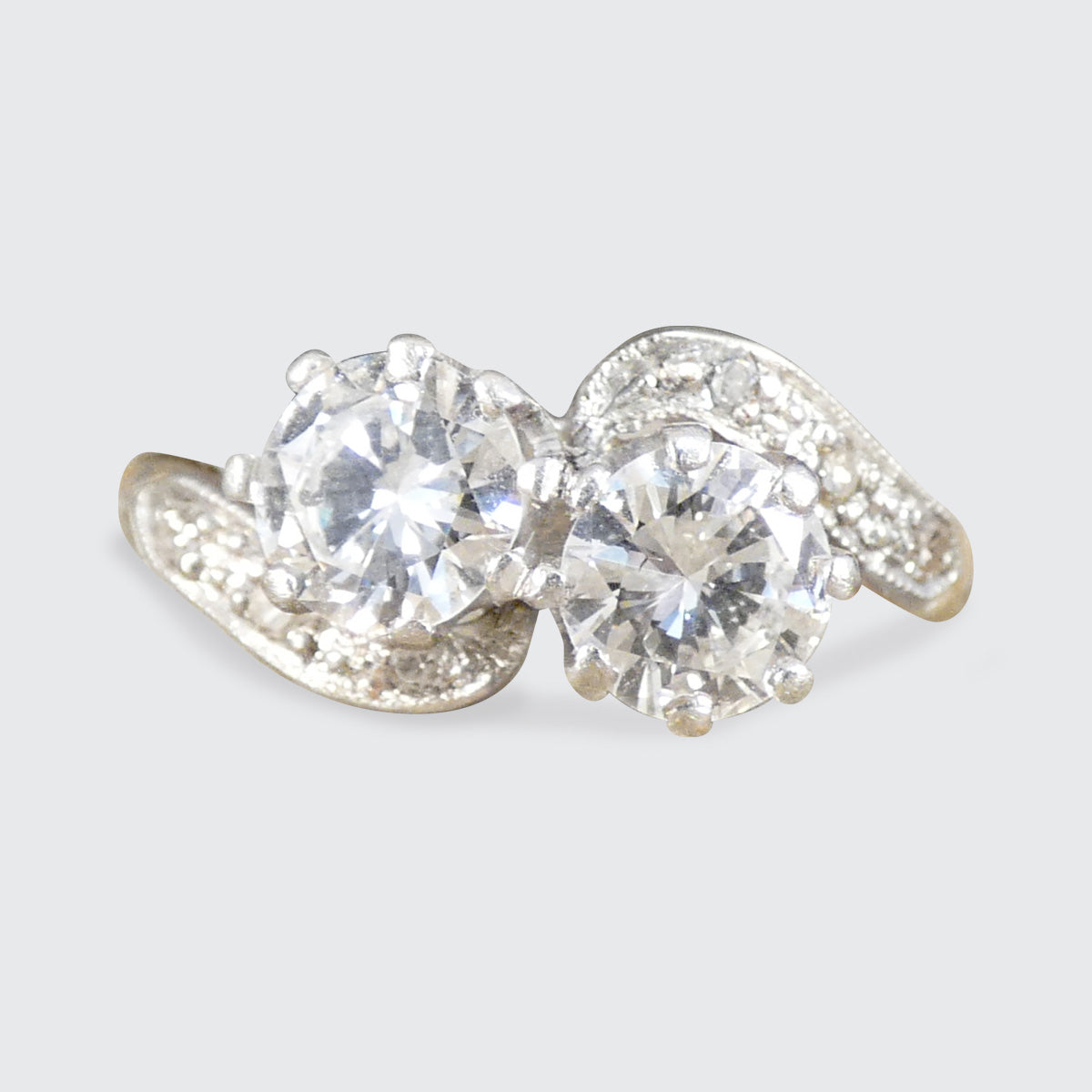 A silver-tone ring featuring two large round clear gemstones in a prong setting, with smaller accent stones along the band, shown on a light gray background.