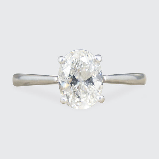 An oval-cut diamond solitaire ring set in a polished silver-colored metal band, shown against a light gray background.