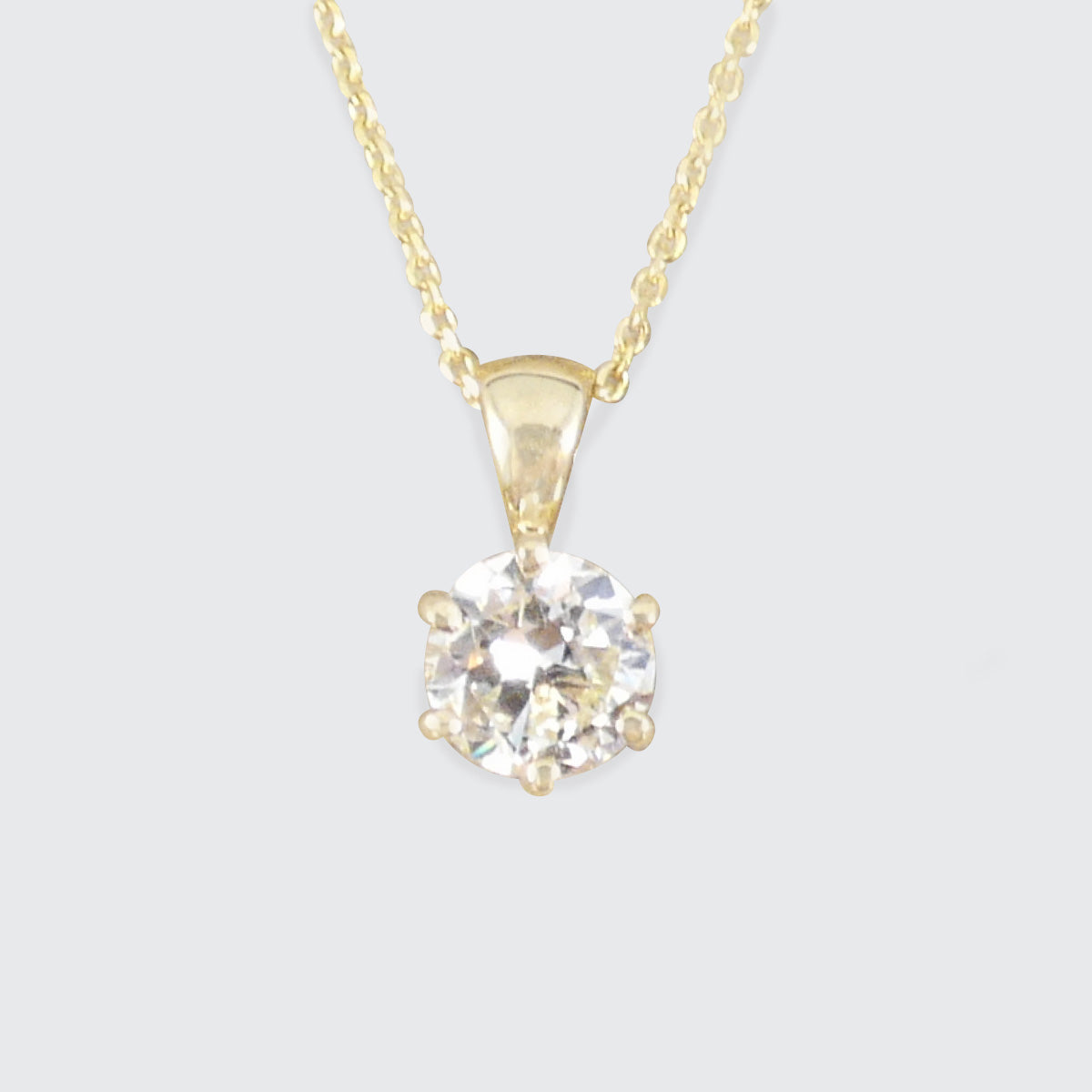 A yellow gold solitaire necklace featuring a round-cut clear diamond pendant in a prong setting, suspended from a delicate gold chain against a light gray background.