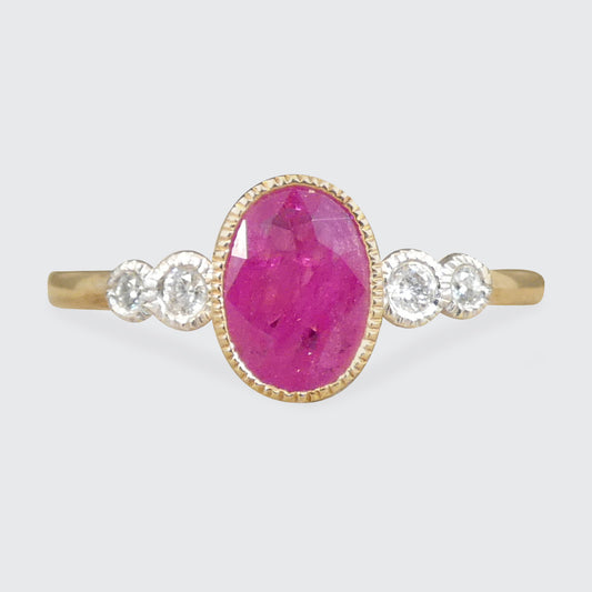 A yellow gold ring featuring a large oval pinkish-red center stone, flanked by three small clear round stones on each side, set against a light gray background.