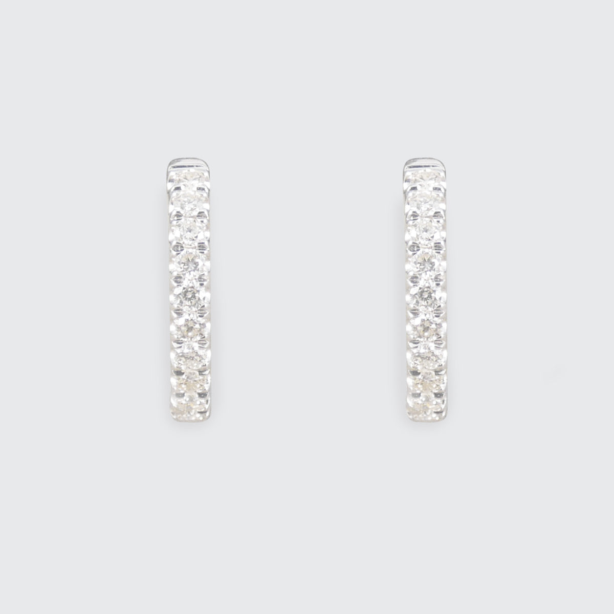 A pair of silver-toned huggie hoop earrings paved with clear cubic zirconia stones, shown against a solid light gray background.