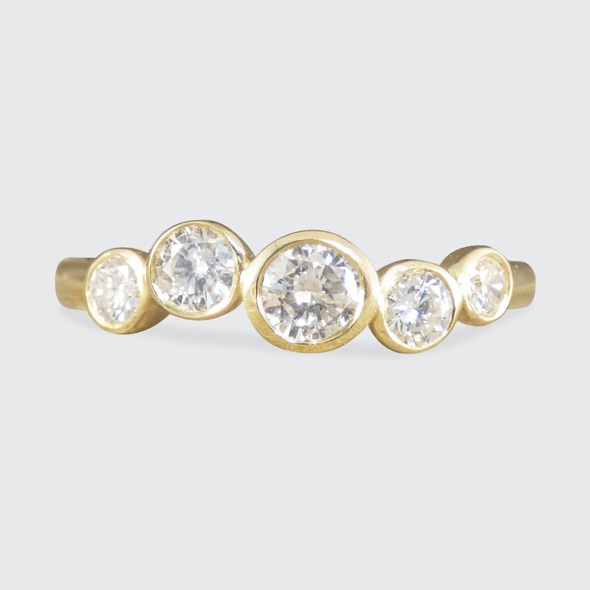 A gold ring featuring five round-cut clear gemstones in bezel settings, arranged in a staggered row against a light gray background.