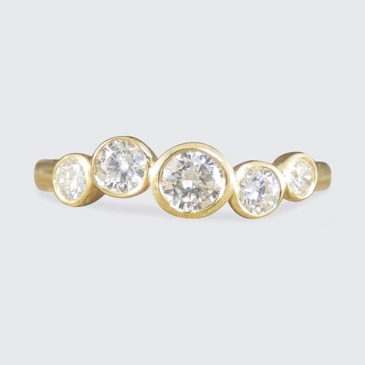 A gold ring featuring five round-cut clear gemstones in bezel settings, arranged in a staggered row against a light gray background.