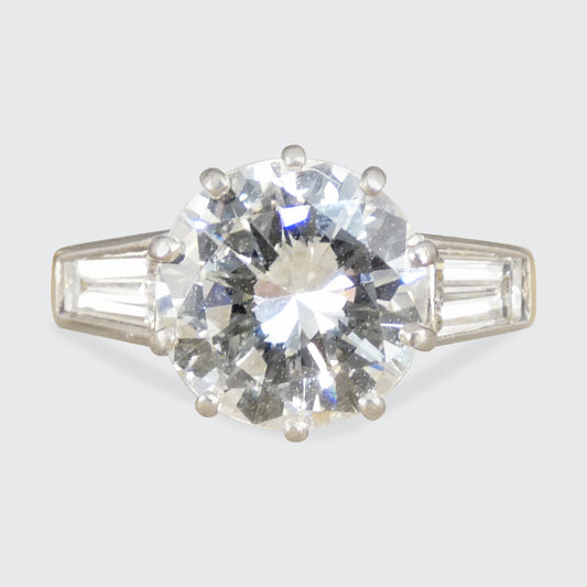 Diamond ring with a large central gemstone and smaller baguette-cut stones on a light gray background