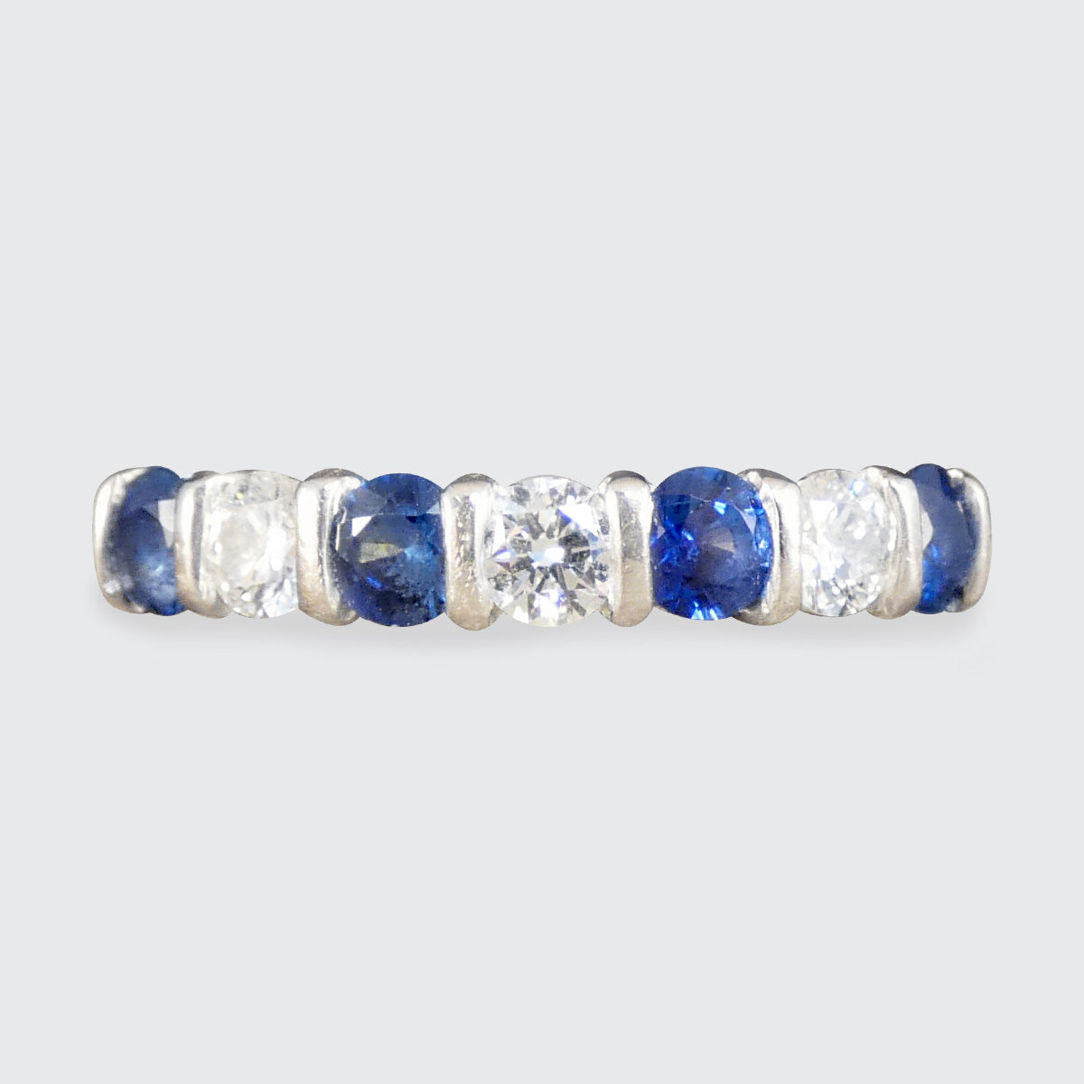 A gold eternity ring featuring alternating round-cut blue sapphires and clear round-cut diamonds, shown against a light gray background.
