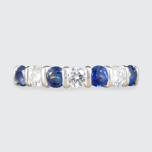 A gold eternity ring featuring alternating round-cut blue sapphires and clear round-cut diamonds, shown against a light gray background.
