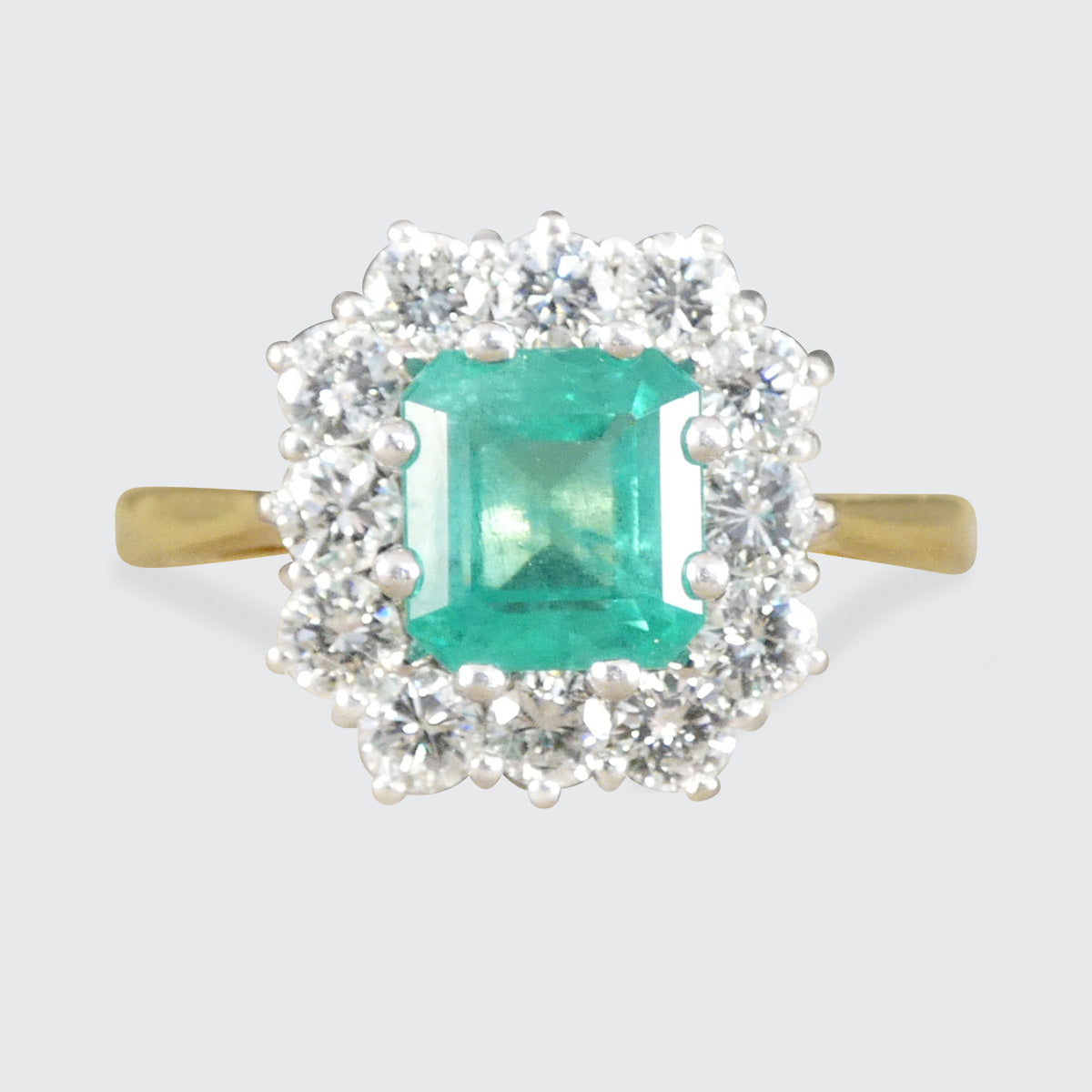 A yellow gold ring featuring a central emerald-cut green gemstone surrounded by a double halo of clear round and baguette-cut stones, set against a light gray background.