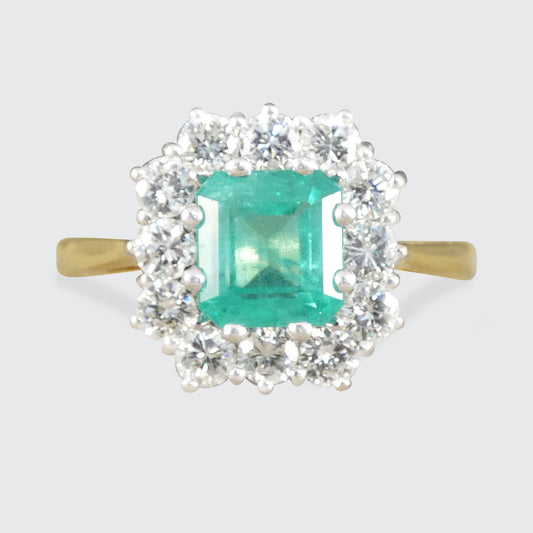 A yellow gold ring featuring a central emerald-cut green gemstone surrounded by a double halo of clear round and baguette-cut stones, set against a light gray background.