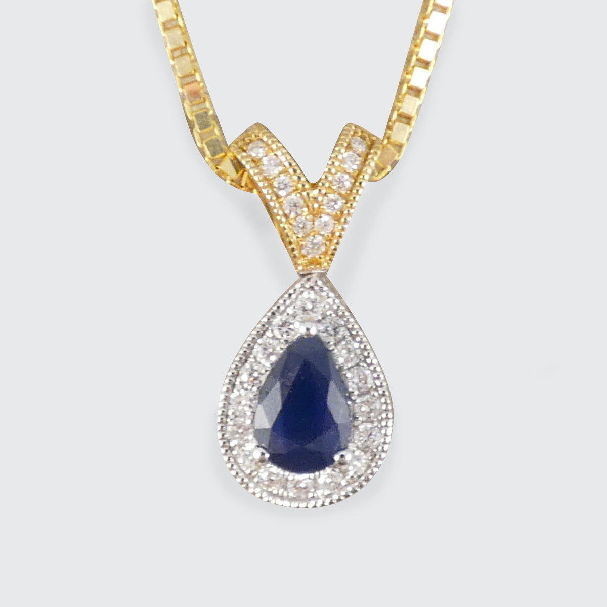 A gold and silver teardrop pendant necklace featuring a deep blue sapphire center stone surrounded by clear pavé diamonds, suspended from a gold box chain against a light gray background.