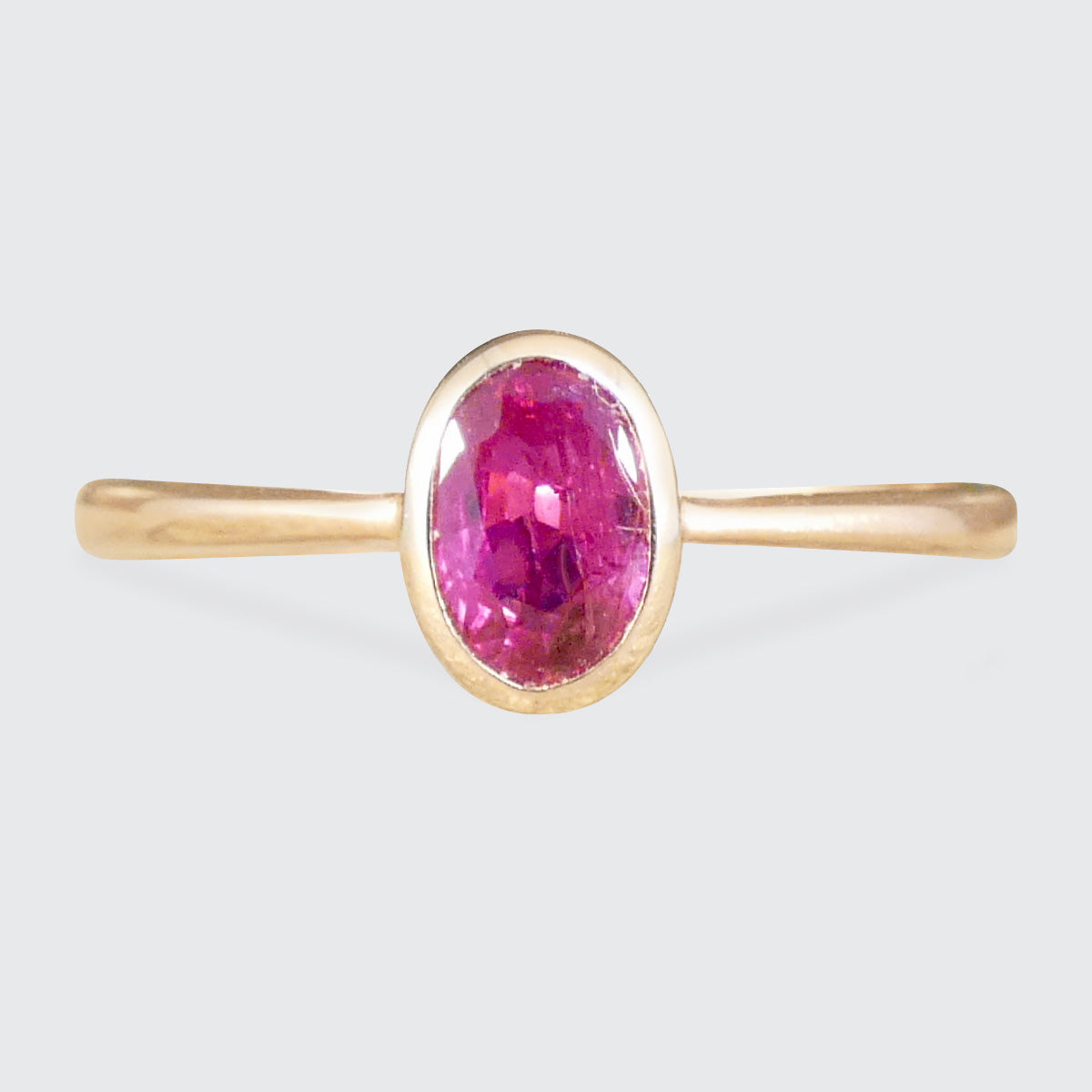 A gold ring featuring an oval-cut pink gemstone in a bezel setting, shown against a light gray background.