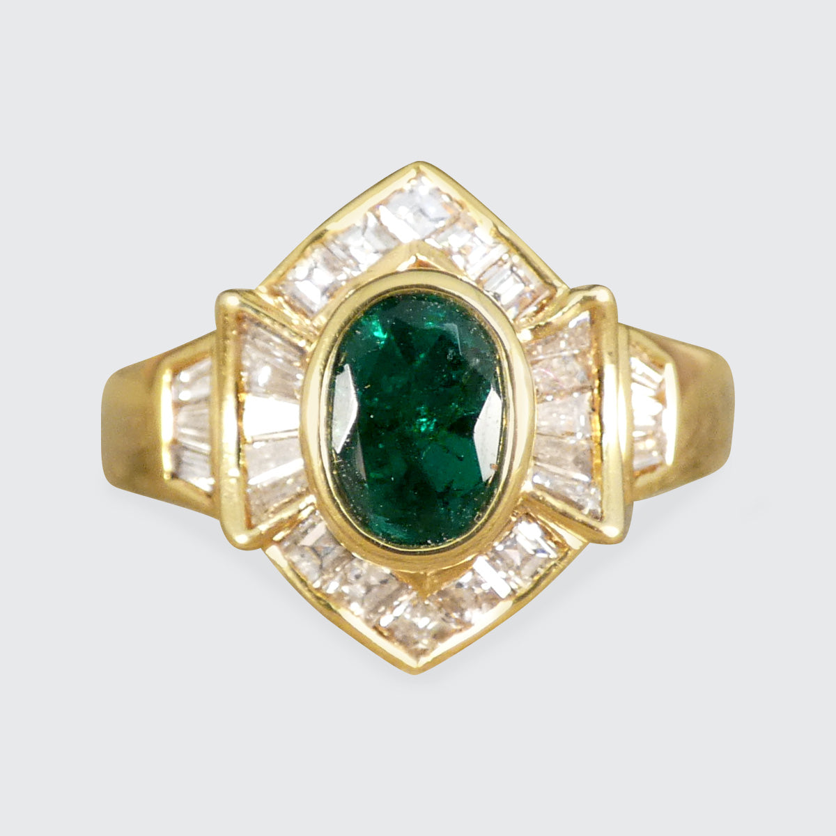 Elegant emerald and tapered diamond cluster ring set in yellow metal, testing as 18ct yellow gold, featuring a vivid oval emerald centre stone surrounded by shimmering tapered baguette diamonds.