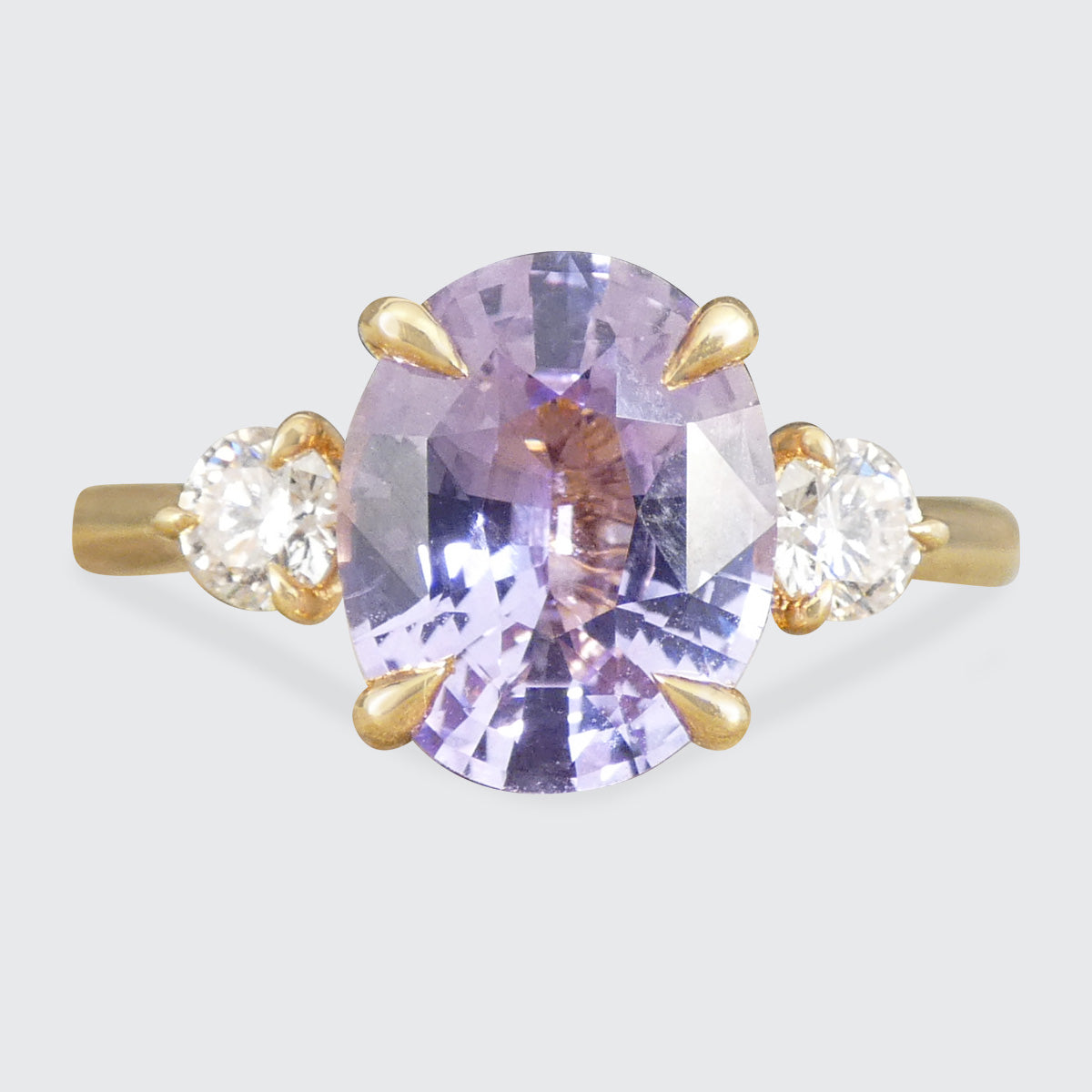 A yellow gold ring featuring a large oval-cut light purple sapphire center stone, flanked by two small round-cut clear diamonds in gold settings, shown against a light gray background.