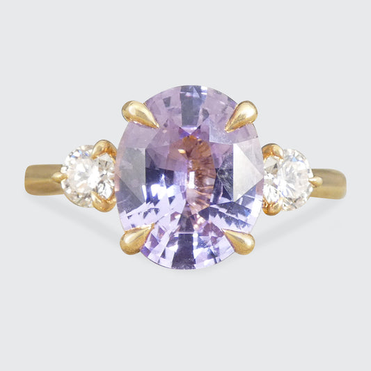 A yellow gold ring featuring a large oval-cut light purple sapphire center stone, flanked by two small round-cut clear diamonds in gold settings, shown against a light gray background.