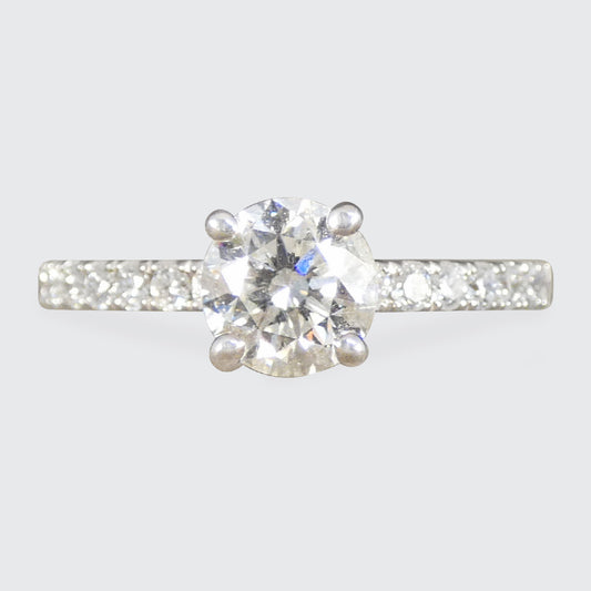 A classic diamond engagement ring with a round-cut center stone in a four-prong setting, featuring a pavé band set with smaller diamonds on a light gray background.