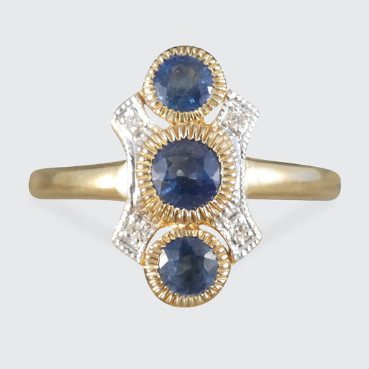 A vintage-style three-stone ring featuring oval blue sapphires and small clear diamonds in a gold and white gold setting, shown against a light gray background.